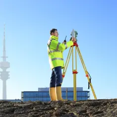 Geodeta w kamizelce odblaskowej obsługuje tachimetr na statywie na tle wieży telewizyjnej i budowy.