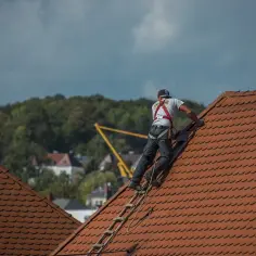 Dekarz w szelkach bezpieczeństwa wspina się po drabinie na czerwony dach.